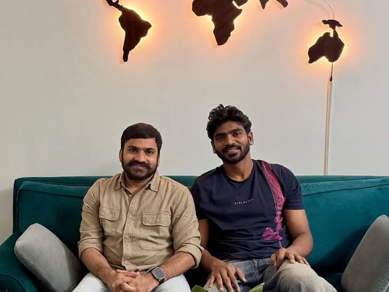 Sandeep Bhaga and cricketer K S Bharat seated on a green sofa in Sandeep's office, beneath a lit world map and his Deputy Commissioner nameplate